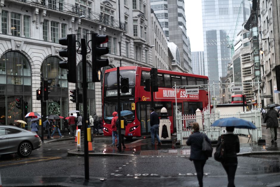 A busy urban street scene during rainy weather in Clapham Junction, featuring a red double-decker bus stopped at a pedestrian crossing. Several pedestrians, some holding umbrellas, are crossing the wet road and waiting at the bus stop. Surrounding the street are tall, modern buildings with large glass windows and ornate architectural details, reflecting the overcast sky. The road surface is shiny with rain, and traffic lights are visible, controlling vehicle and pedestrian flow. Nearby, a gray car is approaching, and other buses can be seen further down the street. The scene captures a typical moment of outdoor city life, with a focus on public transportation, street maintenance, and urban environment, providing an authentic view relevant to outdoor and city landscape themes supported by Gardeners Clapham.