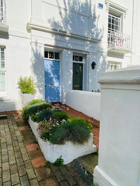 A tidy residential garden in SW4 featuring a narrow, raised flower bed bordered with white painted walls, containing a mix of low-growing green shrubs, pink-flowered plants, and ornamental grasses. Adjacent to the flower bed is a paved pathway made of red bricks and gray cobblestones, leading to a white front door with a blue-painted wooden panel on its side. The exterior of the house is white with large windows, some with white trim and a small balcony above. Shadows from nearby trees cast dappled light onto the garden, highlighting the well-maintained appearance. Gardeners Clapham specialize in garden maintenance, including hedge trimming and lawn care, ensuring the outdoor space remains neat and inviting.