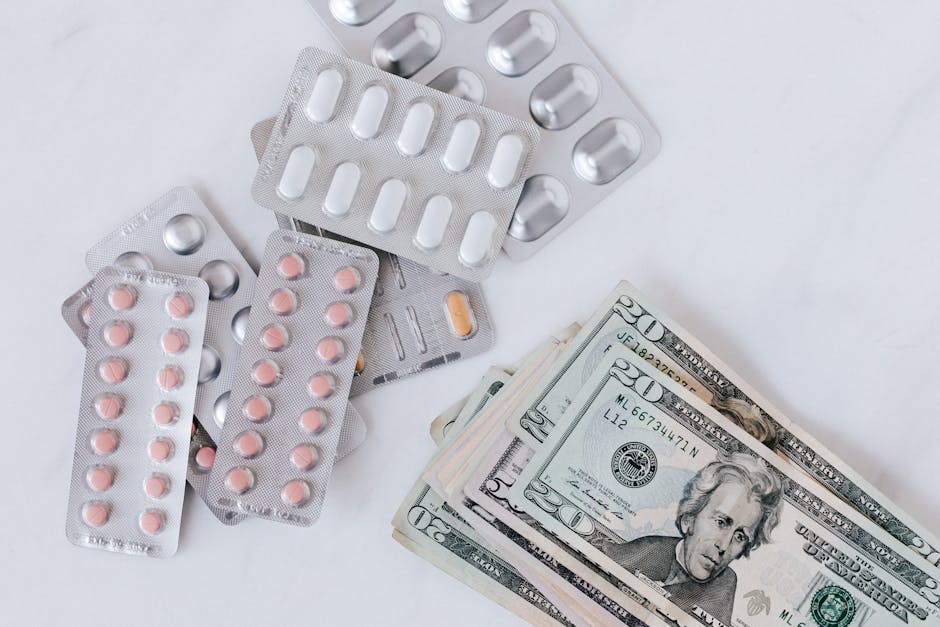An arrangement of various blister packs of pills and medication tablets, including pink and orange capsules, along with several US twenty-dollar bills, are displayed on a plain white surface. The blister packs are slightly overlapping, with some containing small round pills and others empty. The twenty-dollar notes are neatly stacked, showing Benjamin Franklin's portrait, and are positioned towards the lower right side of the image, providing a contrast between medicine and money in a neutral setting. The scene is well-lit, with soft shadows and a clean, uncluttered background, emphasizing the objects for visual clarity. This image may suggest themes related to medical expenses or healthcare costs.