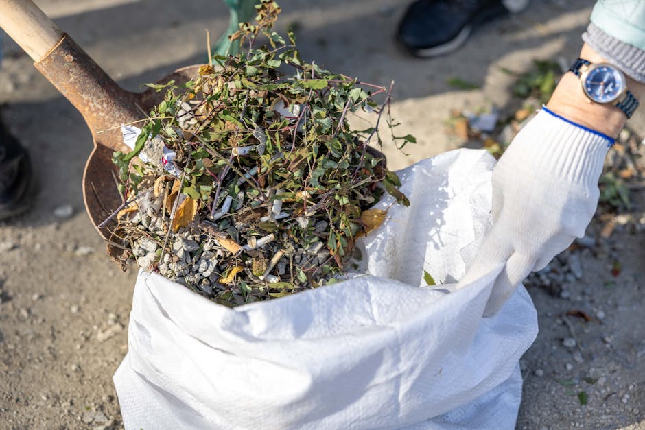 A person wearing a white gardening glove and a wristwatch is seen holding a white garden waste sack filled with a mixture of garden debris, including small twigs, dead leaves, and trimmed branches. A rusty spade is partially visible, positioned over the sack to collect the garden waste. The background shows a gravel or dirt pathway, with the lower part of a person's shoe and some green foliage in the distance. The scene illustrates garden clearance work, emphasizing the collection and disposal of organic waste as part of general garden maintenance. The environment is outdoors, illuminated by natural daylight, with a focus on the tools and debris involved in professional gardening or landscaping tasks, consistent with the services offered by Gardeners Clapham.